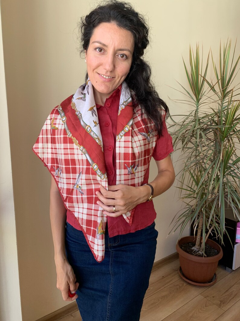 May include: A red and white plaid scarf with a white and gold patterned design. The scarf is tied around the neck of a person wearing a red button-down shirt and blue jeans.