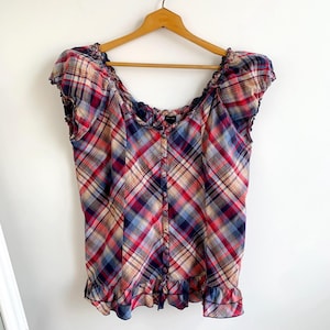 May include: A plaid off-the-shoulder top in red, blue, and beige. The top features a button-down front, ruffled trim along the neckline and hem, and short sleeves. The garment is hanging on a wooden hanger.