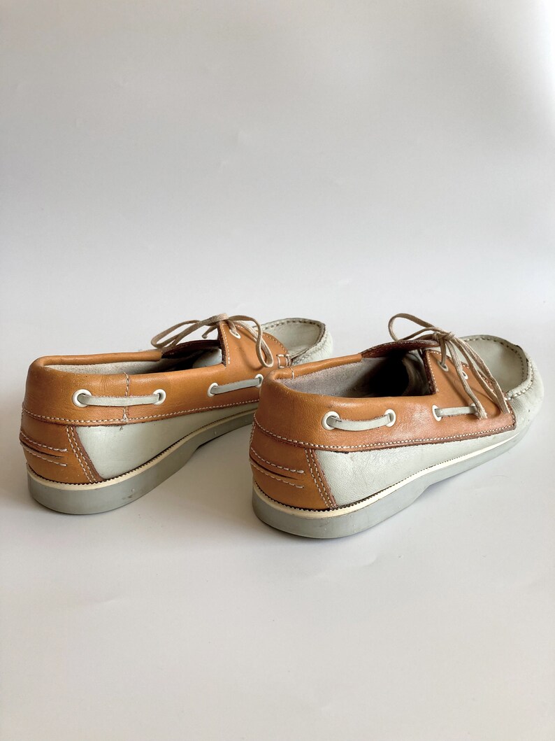 May include: A pair of light grey and brown leather boat shoes with laces. The shoes have a white rubber sole.