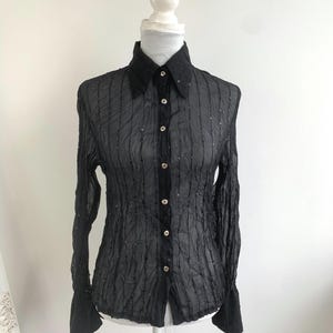 Puede incluir: Camisa negra de manga larga con tejido texturizado y semitransparente. La camisa presenta un cuello clásico, puños abotonados y pequeños adornos decorativos. La camisa se exhibe en un maniquí blanco.