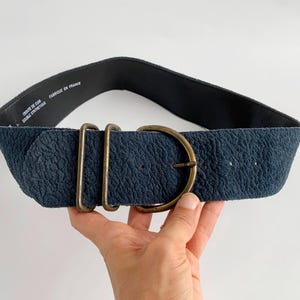May include: A wide, navy blue belt with a textured surface and a large, round, brass-coloured buckle. The belt has two rectangular brass loops next to the buckle. The inside of the belt is black. The text "CUIR DE CUIR DOUBLE SYNTHETIQUE FABRIQUE EN FRANCE" is printed on the inside.