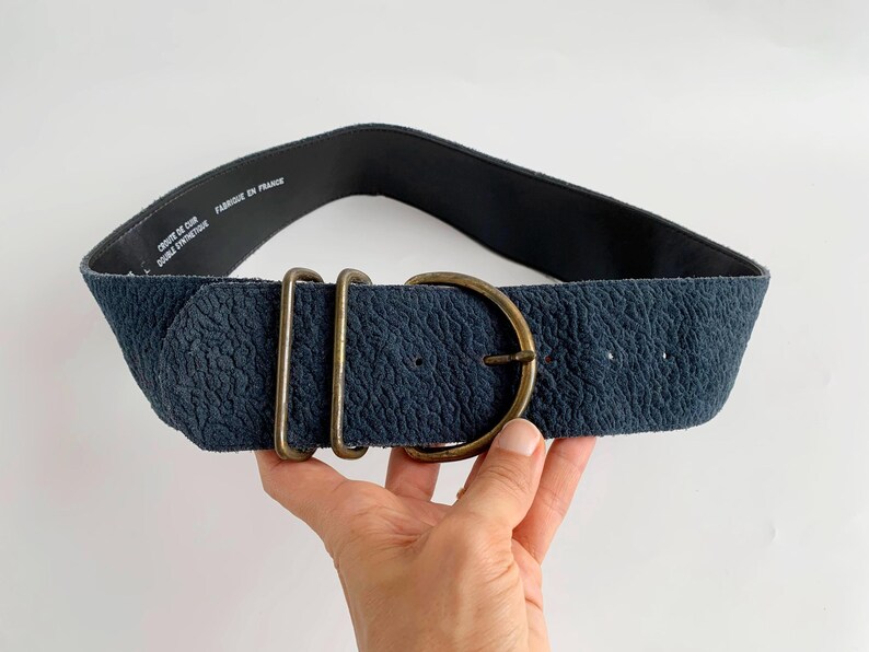 May include: A wide, navy blue belt with a textured surface and a large, round, brass-coloured buckle. The belt has two rectangular brass loops next to the buckle. The inside of the belt is black. The text "CUIR DE CUIR DOUBLE SYNTHETIQUE FABRIQUE EN FRANCE" is printed on the inside.