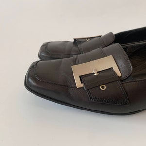 K&ouml;nnte beinhalten: Ein Paar schwarze Leder-Slipper mit einer silbernen Schnalle. Die Slipper haben eine spitze Zehenpartie und einen niedrigen Absatz.