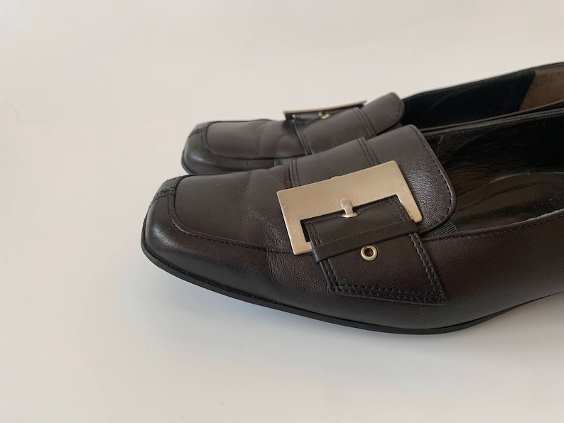 K&ouml;nnte beinhalten: Ein Paar schwarze Leder-Slipper mit einer silbernen Schnalle. Die Slipper haben eine spitze Zehenpartie und einen niedrigen Absatz.