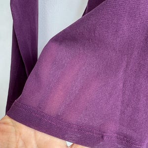 May include: Close-up of a long-sleeved, deep purple garment. The fabric appears to be a lightweight knit, with a subtle sheen. The hem is finished with a neat, double-stitched edge. The sleeve is visible, with a slight transparency.