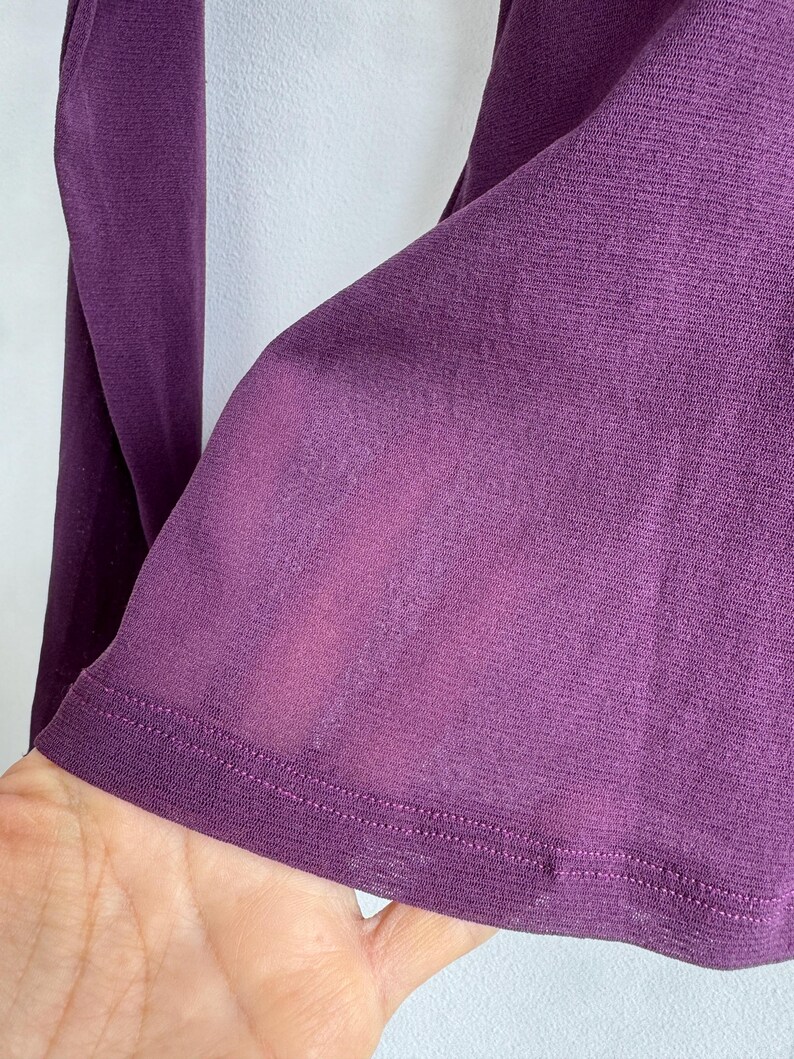 May include: Close-up of a long-sleeved, deep purple garment. The fabric appears to be a lightweight knit, with a subtle sheen. The hem is finished with a neat, double-stitched edge. The sleeve is visible, with a slight transparency.
