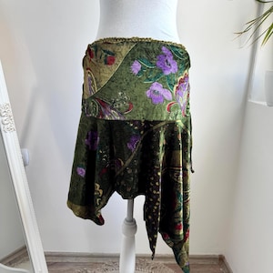 Op de afbeelding: Een knielange rok van groene fluwelen stof met een veelkleurig bloemen- en paisleypatroon in tinten paars, rood en goud. De rok heeft een asymmetrische zoom en een gestructureerde, decoratieve rand langs de tailleband.