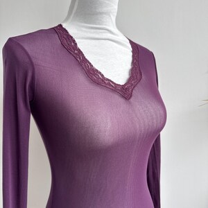 May include: A sheer, long-sleeved, plum-colored top with a V-neckline. The neckline is trimmed with a delicate, dark purple lace. The garment is displayed on a mannequin, showcasing its form-fitting design and lightweight fabric.