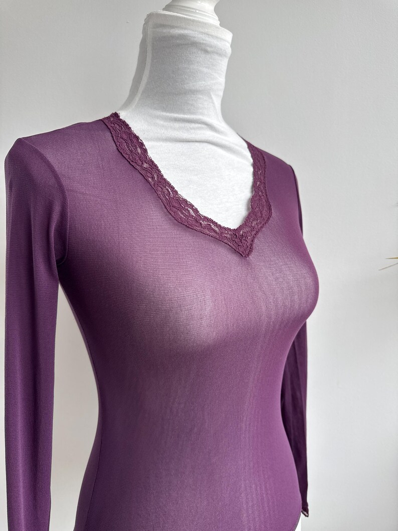 May include: A sheer, long-sleeved, plum-colored top with a V-neckline. The neckline is trimmed with a delicate, dark purple lace. The garment is displayed on a mannequin, showcasing its form-fitting design and lightweight fabric.