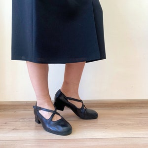 Pu&ograve; includere: Un paio di scarpe in pelle nera con un cinturino sul collo del piede e un piccolo tacco. Le scarpe sono indossate da una persona che indossa una gonna nera.