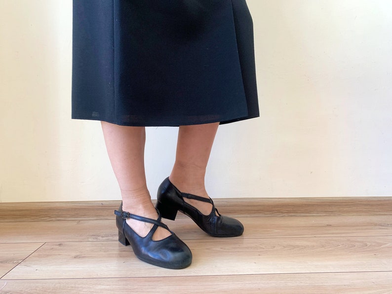 Pu&ograve; includere: Un paio di scarpe in pelle nera con un cinturino sul collo del piede e un piccolo tacco. Le scarpe sono indossate da una persona che indossa una gonna nera.