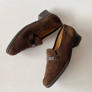 May include: A pair of brown suede loafers with a buckle detail. The loafers have a low heel and a rounded toe.