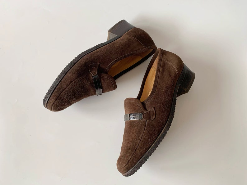 May include: A pair of brown suede loafers with a buckle detail. The loafers have a low heel and a rounded toe.