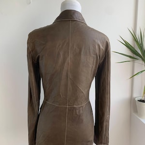 Op de afbeelding: Een bruine leren blazer met lange mouwen en een reverskraag. De jas heeft zichtbare stiksels en een getailleerde pasvorm. De achterkant van de blazer wordt getoond op een mannequin. De jas heeft een neutrale kleur en lijkt van zacht leer te zijn.