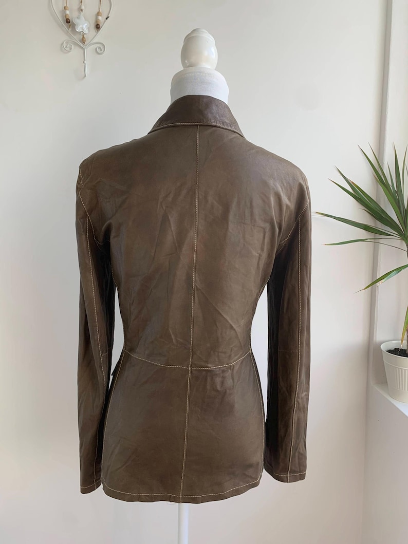 Op de afbeelding: Een bruine leren blazer met lange mouwen en een reverskraag. De jas heeft zichtbare stiksels en een getailleerde pasvorm. De achterkant van de blazer wordt getoond op een mannequin. De jas heeft een neutrale kleur en lijkt van zacht leer te zijn.