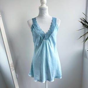 Può includere: Una sottoveste senza maniche color azzurro con un profondo scollo a V decorato da un intricato pizzo in tinta. Il capo è realizzato in un tessuto liscio e satinato che cade morbidamente dal corpetto fino all'orlo, creando una silhouette delicata.