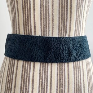May include: A dark teal textured belt is displayed on a mannequin. The wide belt has a crinkled surface and is wrapped around the mannequin's waist. The mannequin is wearing a striped dress.