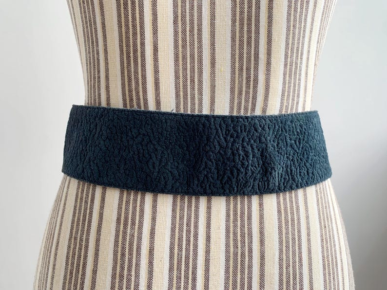May include: A dark teal textured belt is displayed on a mannequin. The wide belt has a crinkled surface and is wrapped around the mannequin's waist. The mannequin is wearing a striped dress.