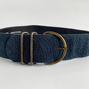 May include: A wide, textured navy blue belt with a large, antique brass buckle. The belt features two rectangular loops and a D-ring buckle. The belt is approximately 7.6 cm wide.