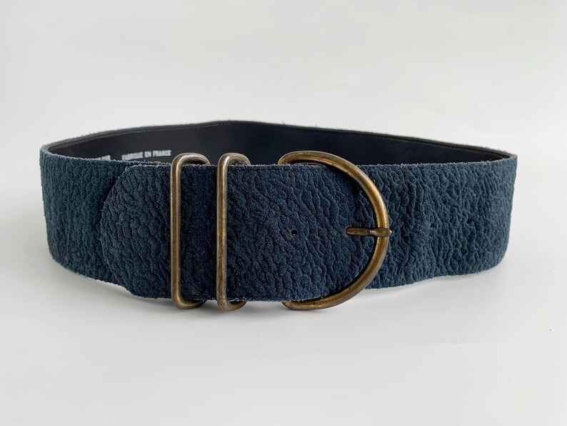 May include: A wide, textured navy blue belt with a large, antique brass buckle. The belt features two rectangular loops and a D-ring buckle. The belt is approximately 7.6 cm wide.