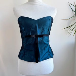May include: A strapless corset-style top in a deep teal, iridescent fabric. The garment features a sweetheart neckline and a vertical black ribbon detail centered on the front, which ties into a small bow at the waistline. The bodice is structured.