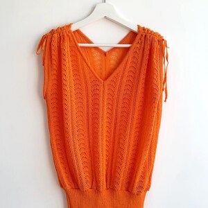 May include: Vibrant orange sleeveless top with a V-neck and an open-knit texture. The top has tie details on the shoulders and a ribbed hem. It is displayed on a white hanger against a plain white backdrop. The knitwear is a striking colour.