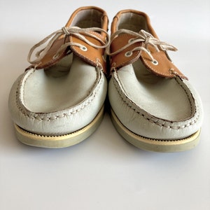 May include: A pair of light brown leather boat shoes with white leather uppers and laces. The shoes have a brown leather trim around the top of the shoe and a white rubber sole.