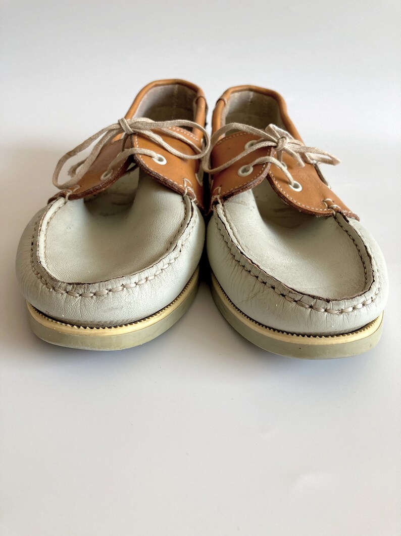 May include: A pair of light brown leather boat shoes with white leather uppers and laces. The shoes have a brown leather trim around the top of the shoe and a white rubber sole.