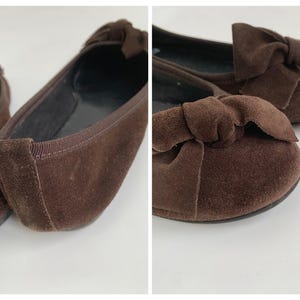 May include: A pair of brown suede ballet flats with a bow detail on the toe. The shoes have a rounded toe and a flat sole. The interior is black. The shoes are displayed on a white surface.