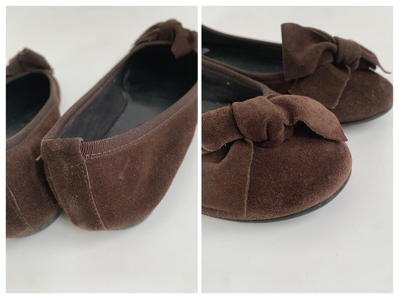 May include: A pair of brown suede ballet flats with a bow detail on the toe. The shoes have a rounded toe and a flat sole. The interior is black. The shoes are displayed on a white surface.