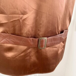 May include: Close-up of a brown satin vest with a leather belt and buckle. The vest has a shiny, smooth texture. The leather belt is a lighter shade of brown and has a rectangular buckle.