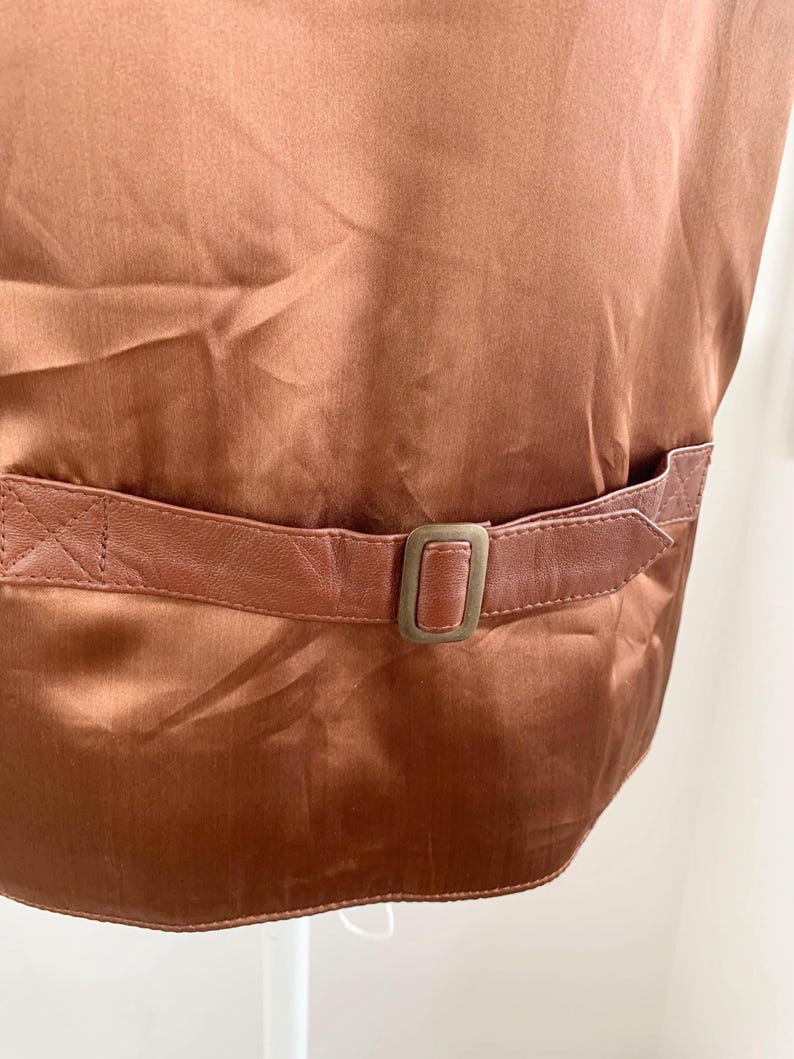 May include: Close-up of a brown satin vest with a leather belt and buckle. The vest has a shiny, smooth texture. The leather belt is a lighter shade of brown and has a rectangular buckle.