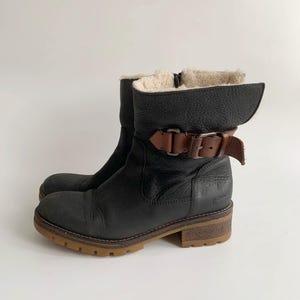 May include: A black leather ankle boot with a textured finish, featuring a brown leather strap with a metal buckle across the ankle. The boot has a cream-coloured faux fur lining, a side zip, and a thick, lugged brown rubber sole.
