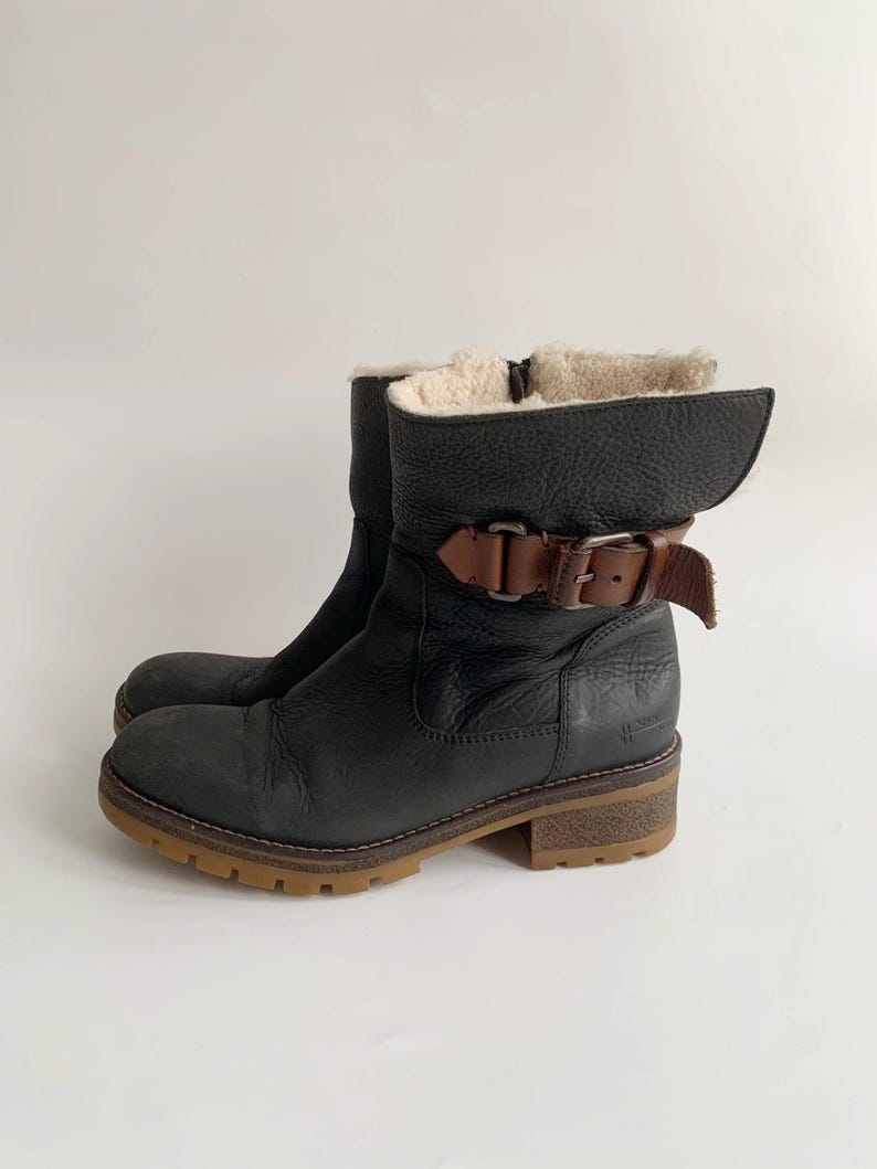 May include: A black leather ankle boot with a textured finish, featuring a brown leather strap with a metal buckle across the ankle. The boot has a cream-coloured faux fur lining, a side zip, and a thick, lugged brown rubber sole.