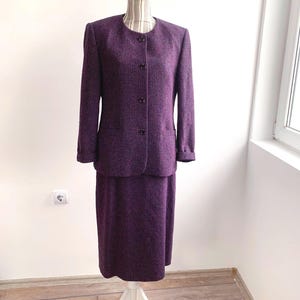 May include: A two-piece purple tweed suit displayed on a mannequin. The suit includes a long-sleeved, button-down jacket with pockets and a knee-length skirt. The fabric has a textured appearance.