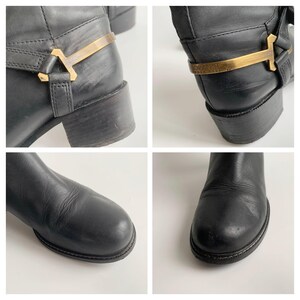 May include: Black leather boots featuring a rounded toe and a low block heel. The ankle area is accented with a decorative gold-tone metal harness strap that includes the embossed text ORANGE HUGO BOSS on the side of the metallic hardware.