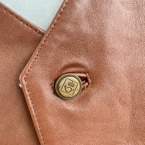 May include: Close-up of a brown leather vest with a single button. The button is round, with a gold-colored metal finish and a decorative emblem. The vest's leather has visible stitching and a slightly textured surface.