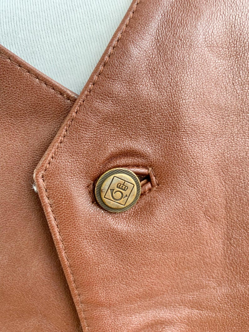 May include: Close-up of a brown leather vest with a single button. The button is round, with a gold-colored metal finish and a decorative emblem. The vest's leather has visible stitching and a slightly textured surface.