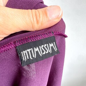 May include: Close-up of a deep purple garment with the brand label "INTIMISSIMI" visible. The label is black with white text and is sewn onto the fabric. The fabric is a sheer material.