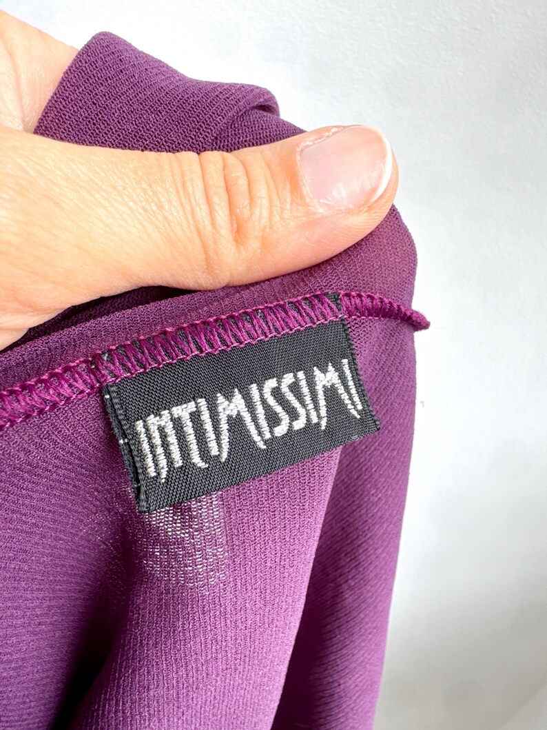May include: Close-up of a deep purple garment with the brand label "INTIMISSIMI" visible. The label is black with white text and is sewn onto the fabric. The fabric is a sheer material.