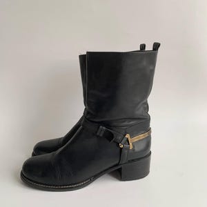 May include: A black leather mid-calf boot featuring a rounded toe, a low block heel, and a decorative strap across the ankle. The strap is secured with a metallic gold-toned hardware accent shaped like a T-bar, positioned on the outer side.