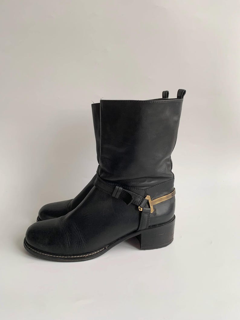 May include: A black leather mid-calf boot featuring a rounded toe, a low block heel, and a decorative strap across the ankle. The strap is secured with a metallic gold-toned hardware accent shaped like a T-bar, positioned on the outer side.