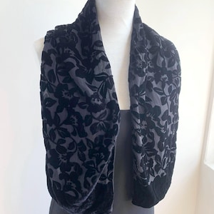 Puede incluir: Una bufanda gris oscuro con un estampado floral. La bufanda está drapeada sobre un maniquí, mostrando su diseño. El estampado floral es de un tono gris más oscuro, creando un contraste sutil. La bufanda parece estar hecha de un material suave y aterciopelado.