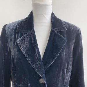 May include: A dark blue velvet blazer with a classic lapel collar and two decorative buttons. The blazer is displayed on a mannequin, showcasing its tailored fit and luxurious texture. The garment has long sleeves and a subtle sheen.