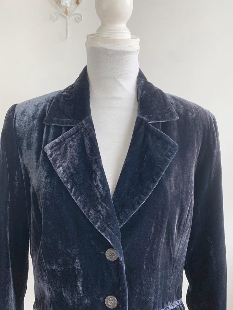 May include: A dark blue velvet blazer with a classic lapel collar and two decorative buttons. The blazer is displayed on a mannequin, showcasing its tailored fit and luxurious texture. The garment has long sleeves and a subtle sheen.