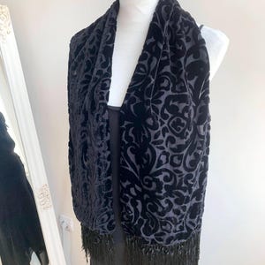 Puede incluir: Bufanda de terciopelo negro con un estampado floral y flecos. La bufanda está drapeada sobre un maniquí, mostrando su diseño y textura. El color oscuro y los detalles intrincados sugieren un accesorio elegante.