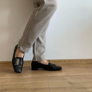 K&ouml;nnte beinhalten: Ein Paar schwarze Leder-Slipper mit silbernen Schnallen. Die Slipper werden zu einer beige Hose getragen.