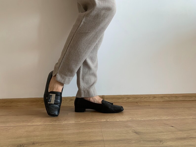 K&ouml;nnte beinhalten: Ein Paar schwarze Leder-Slipper mit silbernen Schnallen. Die Slipper werden zu einer beige Hose getragen.