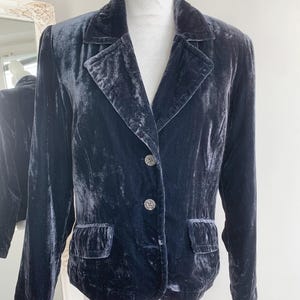 May include: A dark blue velvet blazer with a notched collar, two front pockets, and two decorative buttons. The blazer is displayed on a mannequin. The fabric has a soft, textured appearance.