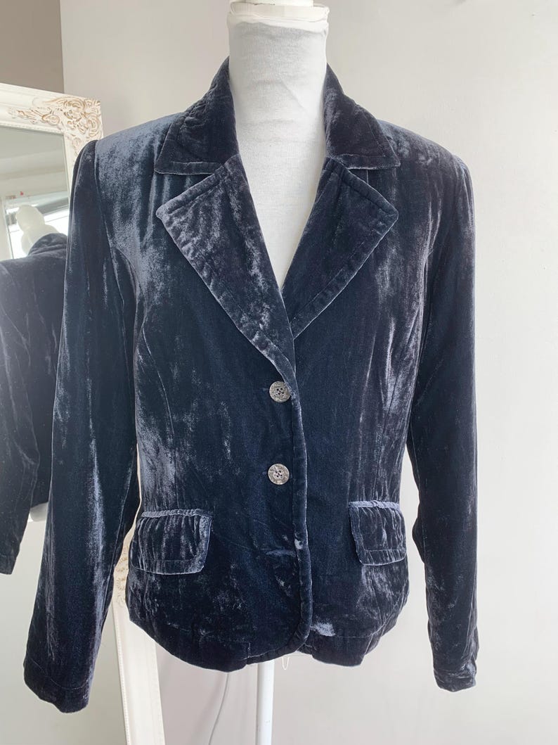 May include: A dark blue velvet blazer with a notched collar, two front pockets, and two decorative buttons. The blazer is displayed on a mannequin. The fabric has a soft, textured appearance.